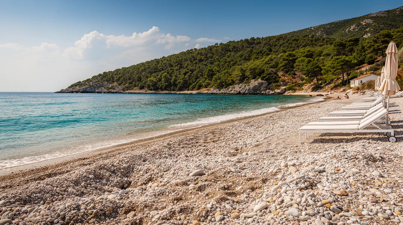 Ein malerischer Kieselstrand an der Ostküste von Korfu, umgeben von einem türkisblauen Meer und einer bewaldeten Küstenlinie, bietet eine entspannende Atmosphäre für Urlauber. Sonnenliegen stehen bereit, um die Sonne zu genießen, während im Hintergrund das charmante Fischerdorf Benitses sichtbar ist.